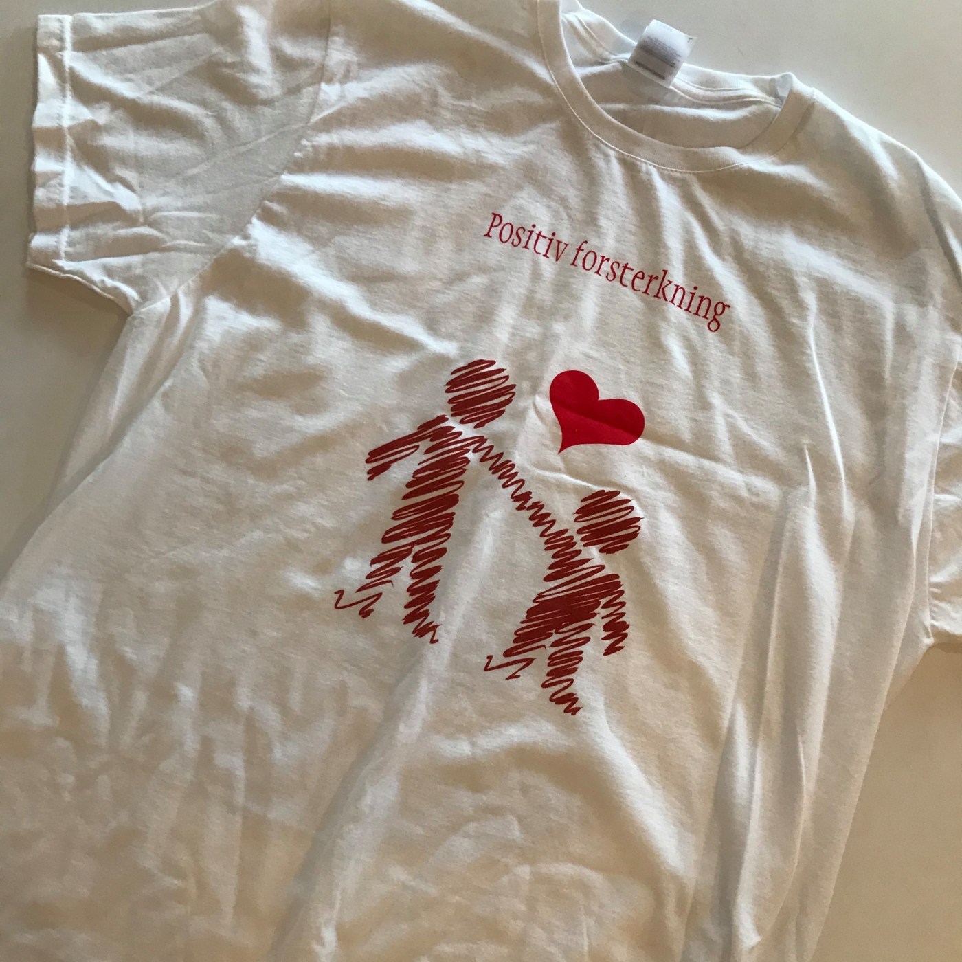 Bilde av t-skjorte med teksten «Positiv forsterkning» og to mennesker som holder hverandre i hånden.